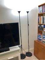 Two identical black metal uplighter floor lamps positioned near a TV stand and bookshelf, showing their size and style.