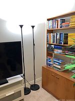 Two black metal torchiere uplighter lamps standing side by side near a TV and bookshelf, showing overall condition and design.