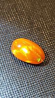 Photo shows a close-up of the large oval cabochon fire opal with bright orange color and visible multicolored flashes on textured black surface.