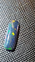 Close-up front view showing multicolored flashes of green, yellow and orange in a long cushion cabochon-shaped black opal on a dark textured background.