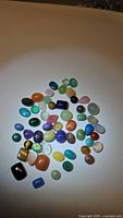 Photo showing a spread of multiple natural polished cabochon gemstones in varied colors and sizes lying on a white surface under lighting.