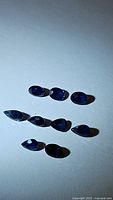 Nine natural blue sapphires laid out on a white surface under varying lighting conditions. Shapes include pear and oval cuts.
