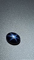 Natural oval cabochon blue star sapphire with 6-ray star effect visible on black textured surface under light.