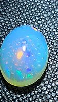Close-up of a translucent oval cabochon cut opal showing blue base color with multicolored flashes including green and orange under light on textured black background.