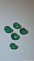 Top view of five natural round cut emeralds arranged individually on a white surface under natural lighting, showing color and clarity.