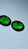 Pair of oval cut electric green tsavorite garnet gemstones under direct light showing their bright green color and internal inclusions.