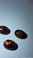 Three polished oval cabochon shaped red tiger's eye gemstones with visible banding and chatoyancy effects on a flat light surface.