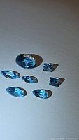 Seven natural Swiss blue topaz gemstones in mixed cuts - oval, square, and marquise shapes displayed on a white surface under light showing color and clarity.