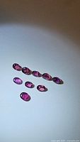 Nine oval cut rubies displayed on white background under light showing pinkish-red color and clarity.