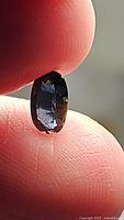Close-up photo of the natural oval cabochon Spinel gemstone held between fingers, showing blue to purple coloration and visible internal characteristics typical of Si1 clarity.