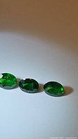 Three oval faceted vivid electric green tsavorite garnet gemstones aligned in a row on a white background, showing their color and clarity.