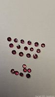 23 small round cut gemstones in varying shades of red to pink-orange arranged on a white background under normal lighting