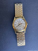 Front full view of the gold-tone men's wristwatch with metal link bracelet on a flat surface.