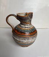 Front-angle view showing the rounded body, wavy spout, and curved handle with horizontal stripes of orange, gray, black, and white