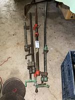 Set of 4 long metal bar clamps with green and red handles, showing wear and rust on concrete floor.