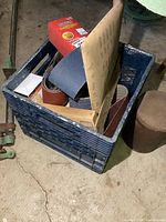 Bin filled with multiple rolls and boxes of sanding belts in various grits and colors, mostly new condition.