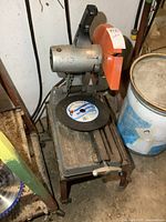 Full view of Makita portable chop saw mounted on metal stand with wheels in workshop corner.