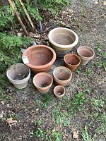 Nine clay pots of various sizes arranged on dirt ground near green foliage. Two pots show visible cracks on rims.