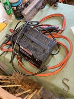 Motomaster battery charger unit with attached power cord and red and black battery clamps lying coiled with heavy-duty orange booster cables on worn green cloth surface.