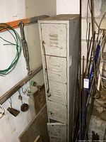 Front view of the tall vintage metal locker with six doors in a storage area. Shows wear, dents, and aged grey paint.