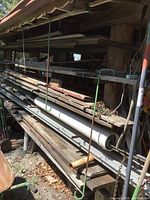 Outdoor rack showing multiple long rough cut lumber pieces stacked horizontally, as well as white and darker metal tubes on the rack