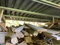 Several older cedar posts and used lumber piled horizontally under a metal roof structure, pieces of plywood partially visible, some cardboard boxes below.