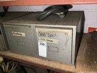 Front view of gray metal 2-drawer cabinet with labels 'Plumbing' and 'Gas Spot Hole Saws' taped onto the drawers showing wear and aging.