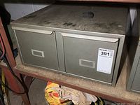 Front view of metal 2-drawer cabinet with seller tag visible, showing cabinet on shelf with dust on surface.