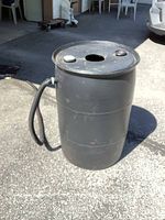 Full view of the black plastic rain barrel standing upright on pavement, hose attached to the side.