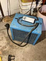 Blue electric shop heater with metal grille and attached power cord and plug, photographed from front angle on concrete floor in workshop setting.