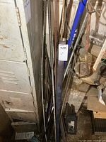 Multiple steel threaded rods leaning against a wall next to a white locker and some tools. Different sizes of rods visible, with some showing threaded ends.