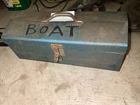 Metal toolbox closed with 'BOAT' written on top, showing surface wear and rust spots, secured with a latch and featuring a metal handle.