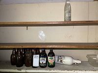 Several vintage bottles arranged on shelves, including 5 brown stubby bottles and two labeled colored bottles with a white stoneware bottle lying sideways.