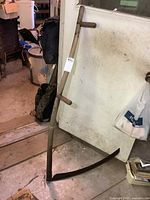 Full view of the entire vintage scythe leaning against a door indoors, showing wooden handle and metal blade.