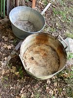 Two old metal washtubs placed outdoors on the ground showing rust, dirt, and dents.