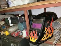 Welding helmet with flame graphics and several tubes of welding rods on a shelf.