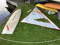 Two white windsurfing boards on grass by water with one sail laid next to them.