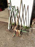 Front view of the yard tools group including axe, pitchforks, rakes, shovels, and hoes leaning against a white wall outside