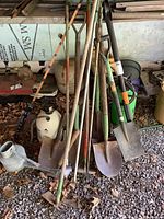 Full view of all yard tools grouped together on gravel floor showing shovels, hoe, watering cans, and sprayers.