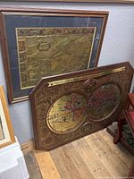 Two gold-toned world atlas wall hangings leaning against a wall, one framed and one decorative board with two circular maps.