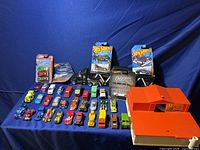 Photo showing the collection of approximately 35 die-cast toy cars in various colors, a black toy car carrier truck, two Hot Wheels cars in packaging, and the orange vintage playset.