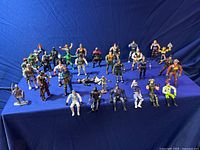 Full view of approximately 35 mini vintage action figures arranged in rows on blue fabric background, showing color and poses.
