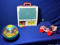 Three vintage Fisher Price toys displayed on a blue cloth backdrop: chalkboard desk toy, pull toy, and chime ball.