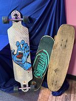 Three long skateboards displayed upright and stacked, showing a colorful graphic skateboard with a blue screaming hand, a black and green Powell Peralta skateboard, and a plain wooden deck.