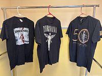 Three black rock band T-shirts hanging on rack featuring Linkin Park, Nirvana, and DEVO graphics