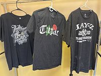 Three black T-shirts hanging on a rack; Tupac, Ice Cube, and Jay-Z themed designs visible.