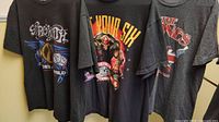 Three rock band T-shirts hung side by side showing front printed designs for Rolling Stones, Five Finger Death Punch, and Aerosmith.