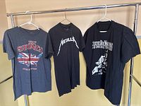 Three rock band T-shirts hanging on a rod, showing The Rolling Stones, Metallica, and Stevie Ray Vaughan and Double Trouble shirts.