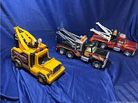 Three vintage toy tow trucks including two Nylint (one yellow, one black) and one maroon Fisher Price, set against a blue cloth background.