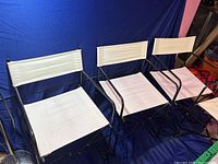 Four white and black folding chairs lined up against a blue cloth backdrop. Chairs have black tubular metal frames and white fabric seating surfaces.